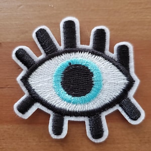 May include: An embroidered eye patch in white and blue with black eyelashes. The eye is surrounded by a white circle with a blue outline. The eyelashes are black and extend out from the eye.