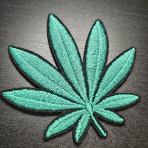 May include: Embroidered patch of a teal cannabis leaf with black edging. The patch has detailed stitching, showing the leaf's veins. Suitable for clothing, bags, or other fabric items. The patch is approximately 7 cm in length.