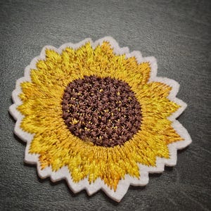May include: Embroidered sunflower patch. The design features a yellow and brown sunflower with a white border. The patch is detailed and textured, suitable for application on clothing or accessories.