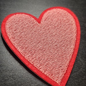 May include: A red heart-shaped fabric patch with a textured, woven centre and a solid red border. The patch is designed to be sewn or ironed onto clothing or other items. The heart shape is a classic symbol of love and affection.