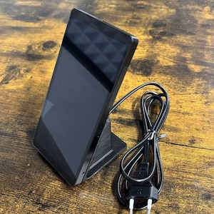 Op de afbeelding: Een zwart rechthoekig elektronisch apparaat met een donker scherm staat rechtop op een zwarte 3D-geprinte standaard. Een zwarte stroomkabel met een tweepolige stekker ligt opgerold naast de standaard. Het apparaat heeft een glad, donker frame.