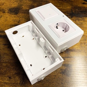 Op de afbeelding: Een witte rechthoekige elektrische wanddoos met bijpassende afdekplaat voorzien van een lichtschakelaar en een geaard stopcontact. De unit wordt getoond met de holle montagebasis losgekoppeld, waardoor interne schroefgaten zichtbaar zijn.