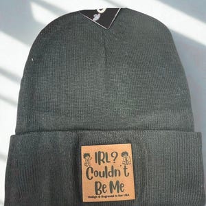 May include: Dark grey beanie hat with a turned-up brim. A square, tan leather patch is sewn on the front, with the text "IRL? Couldn't Be Me" and small cartoon figures. The text "Design & Engraved in the USA" is printed below.