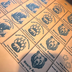 May include: A collection of square, tan leather patches with various bear-themed designs and text. The patches feature images of bears, bear paws, and phrases like "Bearly Awake" and "Great Smoky Mountains." Each patch is designed and engraved in the USA.