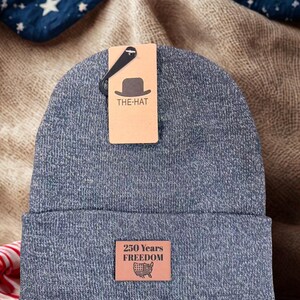 May include: A blue beanie with a folded brim. A tag reads "THE-HAT" with a hat graphic. A leather patch displays "250 Years Freedom" and a map of the United States. The hat is displayed on a textured surface.