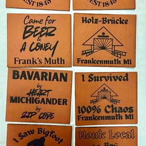 May include: A collection of brown leather coasters with various black text designs. Some coasters feature the text "Frankenmuth MI EST 1845", while others have phrases like "Came for Beer & A Coney" and "I Saw Bigfoot".