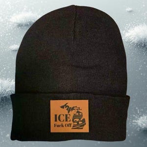 Op de afbeelding: Zwarte beanie met een bruine leren patch. De patch toont de omtrek van Michigan met de woorden "ICE" en "FUCK OFF" in een vetgedrukte lettertype. De beanie is gemaakt van een zacht, gebreid materiaal.