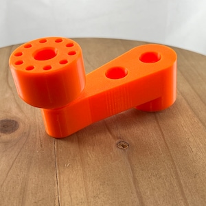 Könnte beinhalten: Ein orangefarbenes Plastikwerkzeug mit einem runden Griff und einem langen Arm mit zwei Löchern. Der Griff hat eine Reihe kleiner Löcher am Rand.