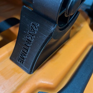 Pu&ograve; includere: Primo piano di un attacco per passeggino in plastica nera con la scritta "ULTIMATE" stampata su di esso. L'attacco &egrave; fissato a un passeggino con una maniglia nera e un telaio arancione.