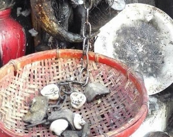 Op de afbeelding: Een donker, bijna zwart figuur met een mensachtig gezicht is bedekt met spijkers, botten en andere voorwerpen. Een ketting hangt van het figuur en verbindt met een geweven mand gevuld met witte voorwerpen. Het figuur is omringd door andere voorwerpen, waaronder een rode kalebas, een bruin hoorn en een witte plaat.