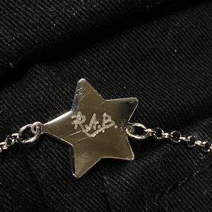 Puede incluir: Un charm plateado en forma de estrella con las iniciales "R.A.B." grabadas en él, unido a una cadena plateada.
