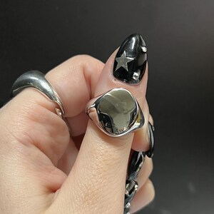 Può includere: Un anello a sigillo d'argento con una parte superiore liscia a forma ovale. L'anello è indossato su un dito con smalto nero e nail art a stella argentata.