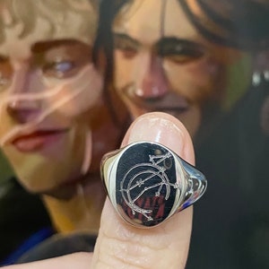 May include: A silver signet ring with an oval face featuring a detailed engraved design of a crescent moon and a stylized star with a barbed wire border.