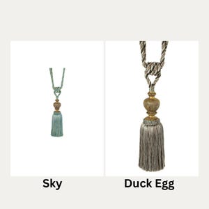 May include: Two decorative curtain tiebacks with tassels. The tieback on the left is sky blue with a gold bead. The tieback on the right is a light grey with a gold bead.