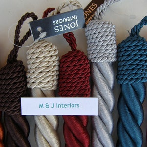 May include: Assortment of twisted rope curtain tiebacks in various colours, including orange, brown, beige, red, silver, teal, and pink. The tiebacks are made of a soft, woven fabric and feature a decorative knot at the end. The tiebacks are labelled with the text "M & J Interiors" and "Jones Interiors".