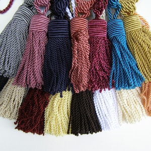 May include: A collection of decorative tassels in various colors, including black, gray, beige, burgundy, navy, pink, teal, gold, and white. Each tassel features a twisted rope design and a looped top for hanging. These tassels are suitable for home decor and craft projects.