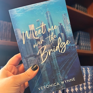 May include: A book cover with a blue and white cityscape background. The title of the book is "Meet me over the Bridge" in gold lettering. The author's name, "Veronica Wynne", is printed in white at the bottom of the cover.
