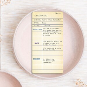 May include: A vintage-style library card menu for Sam's 30th Birthday, featuring appetizers, main courses, and dessert options. The menu includes terrine, slow-roasted chicken, wild mushroom risotto, and key lime pie. The card is set on a pink plate.