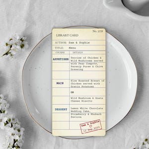 以下が含まれることがあります： 「LIBRARY CARD」と「Menu」の文字が入ったヴィンテージスタイルのメニューカード。前菜、メインコース、デザートが記載されています。カードは金色の縁取りのある白いお皿の上にあり、背景には白い花があります。