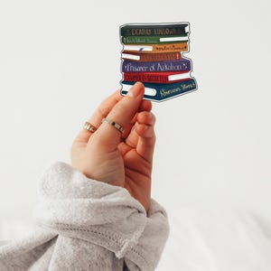 Op de afbeelding: Een sticker met een stapel kleurrijke boeken met titels als "Deathly Hallows" en "Sorcerer's Stone". De boeken zijn in zwart, groen, bruin, paars, rood en blauw. De sticker staat tegen een witte achtergrond.