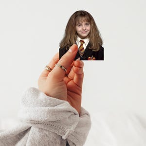 May include: A small rectangular image of a person with long brown hair, wearing a black blazer and a striped tie. The image is held by a hand wearing two rings, against a white background.