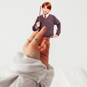 May include: A cutout of a person with red hair, wearing a gray sweater, a white collared shirt, and a tie, holding a wand. The person is being held up by a hand wearing rings, with a gray sweatshirt sleeve in the foreground.