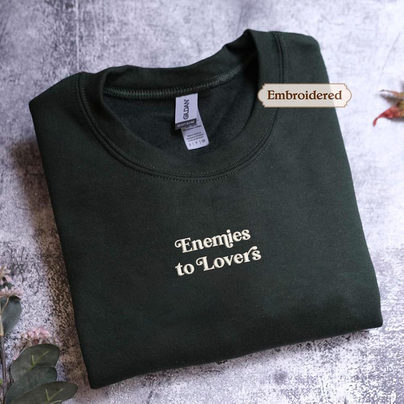 A dark green sweatshirt with the text " Enemies to Lovers" embroidered on it, lying on a textured surface.