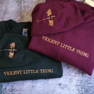 May include: Two sweatshirts, one dark green and one burgundy, are folded and stacked. Each has a gold embroidered dragon design and the words "VIOLENT LITTLE THING." The burgundy sweatshirt is on top, with the text visible. A book and leaves are in the background.