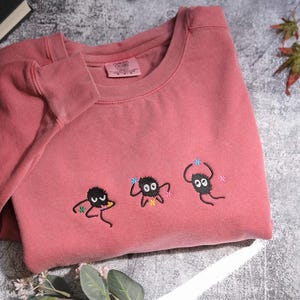 a pink t-shirt with three small black creatures with white eyes and antennae, as well as some leaves, placed on a gray surface.