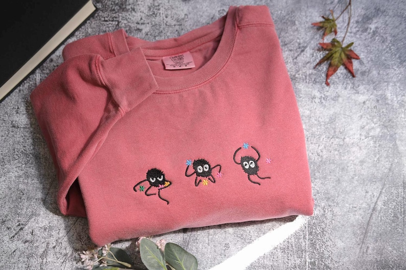 a pink t-shirt with three small black creatures with white eyes and antennae, as well as some leaves, placed on a gray surface.