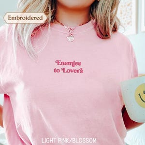 A person wearing a pink t-shirt with the text "Enemies to Lovers" embroidered on it, holding a white mug with a smiley face on it.