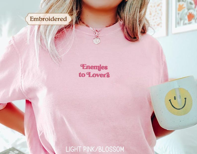 A person wearing a pink t-shirt with the text "Enemies to Lovers" embroidered on it, holding a white mug with a smiley face on it.