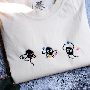 a white t-shirt with three small black figures with colorful accents, each holding a star, arranged in a row.