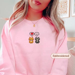 A woman wearing a pink sweater with a pixel art design of two cats and a heart symbol.