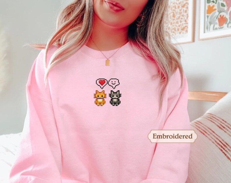 A woman wearing a pink sweater with a pixel art design of two cats and a heart symbol.