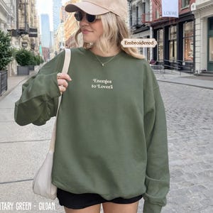 A woman wearing a green sweatshirt with the text "Enemies to Lovers" printed on it, standing on a city street.
