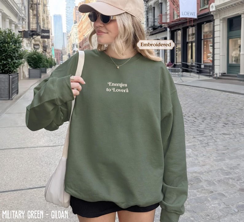 A woman wearing a green sweatshirt with the text "Enemies to Lovers" printed on it, standing on a city street.