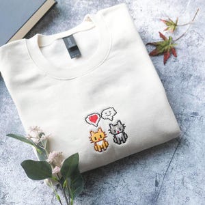a white t-shirt with a pixel art design of two cats and a heart, along with a small plant and some leaves.