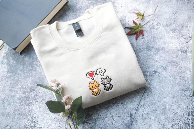 a white t-shirt with a pixel art design of two cats and a heart, along with a small plant and some leaves.