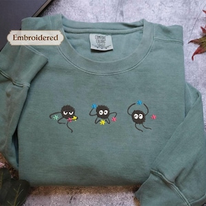 a green long-sleeved shirt with three small black creatures with white eyes and colorful accents, such as flowers, on the front. The shirt is placed on a gray surface, and there are some leaves and a plant visible in the background.