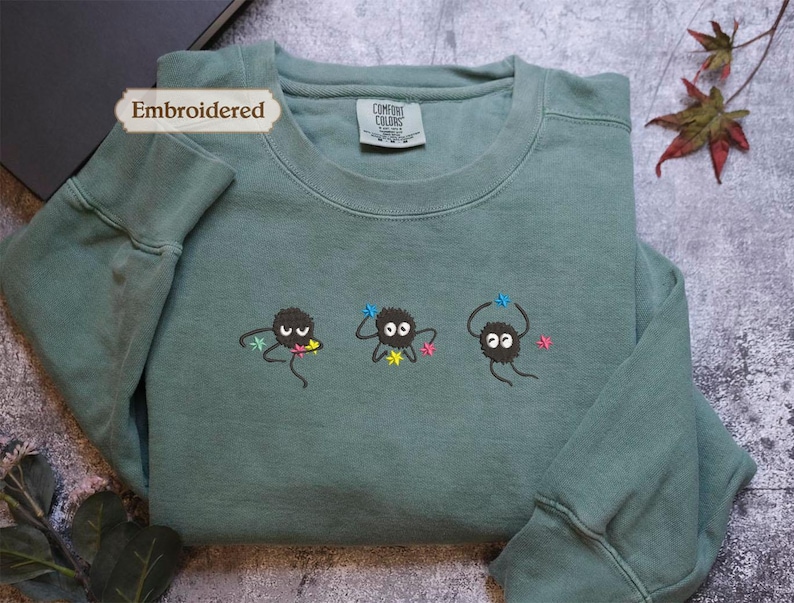 a green long-sleeved shirt with three small black creatures with white eyes and colorful accents, such as flowers, on the front. The shirt is placed on a gray surface, and there are some leaves and a plant visible in the background.