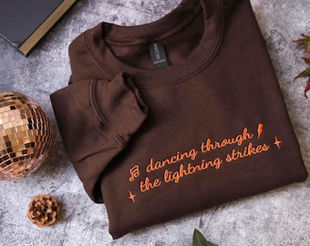 May include: A dark brown crewneck sweatshirt featuring orange embroidered text that reads "dancing through the lightning strikes" accompanied by a musical note, a lightning bolt, and two small four-pointed star icons on the front chest area.
