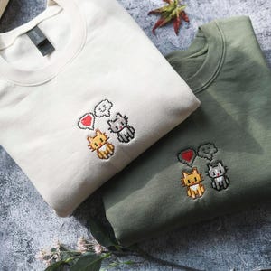 two t-shirts with cat designs, one white and one green, laid out on a surface with some books and leaves.