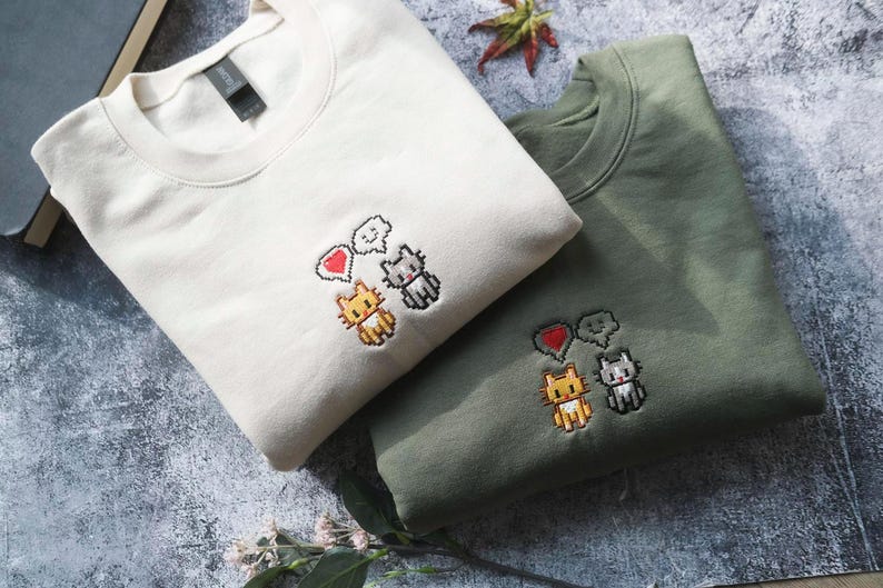 two t-shirts with cat designs, one white and one green, laid out on a surface with some books and leaves.
