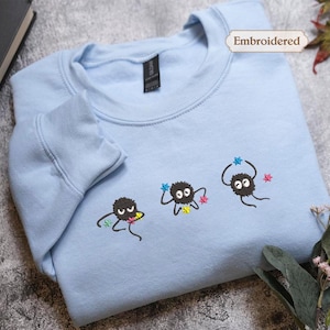 a light blue t-shirt with three small black creatures with googly eyes and colorful arms, along with a label that says "Embroidered". The t-shirt is placed on a gray surface, and there is a small plant and a book visible in the background.