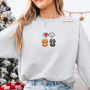 A woman wearing a gray sweatshirt with a pixelated design of two cats and a heart symbol.