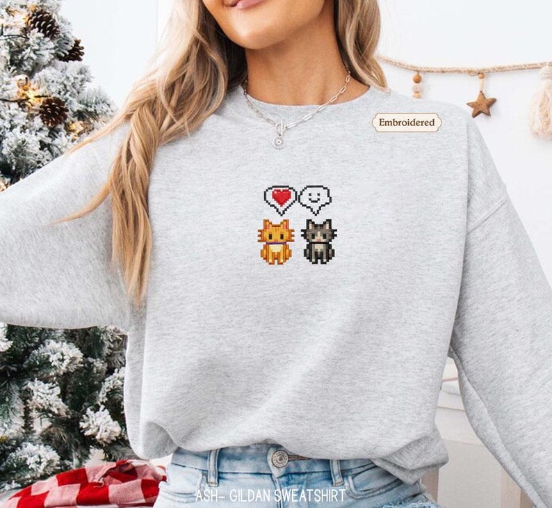 A woman wearing a gray sweatshirt with a pixelated design of two cats and a heart symbol.
