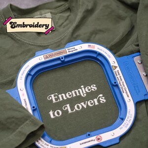 A green t-shirt with a blue embroidery frame that has the text "Enemies to Lover's" printed on it.