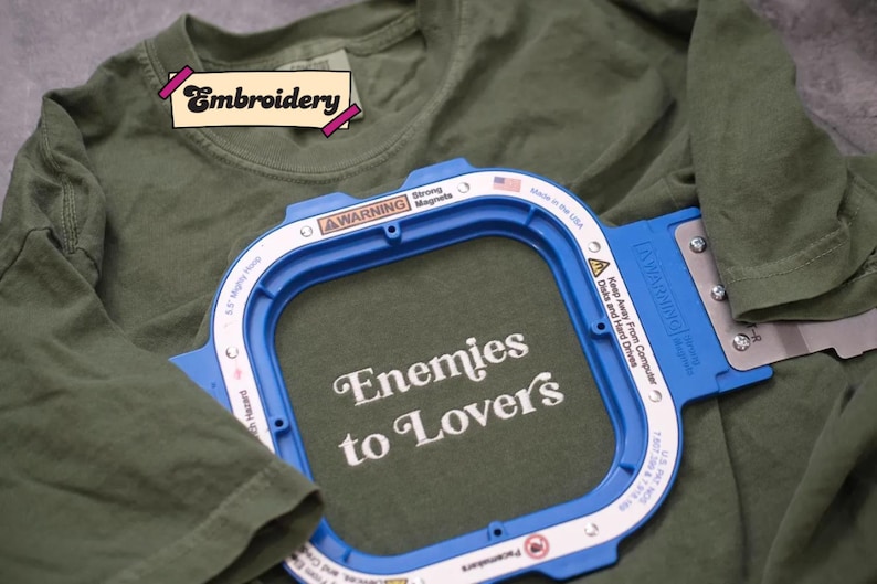 A green t-shirt with a blue embroidery frame that has the text "Enemies to Lover's" printed on it.