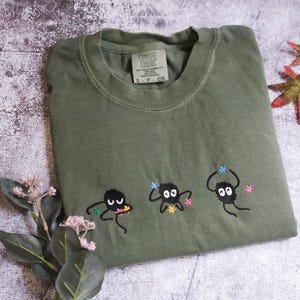 a green t-shirt with three small black figures and flowers embroidered on it, along with some leaves and flowers on a textured surface.